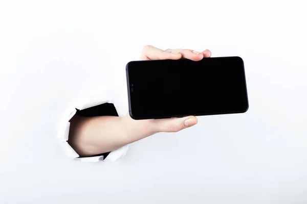 Mano femenina fuera del agujero en el papelero, sosteniendo un teléfono inteligente con una pantalla negra. Aislar sobre fondo blanco . —  Fotos de Stock