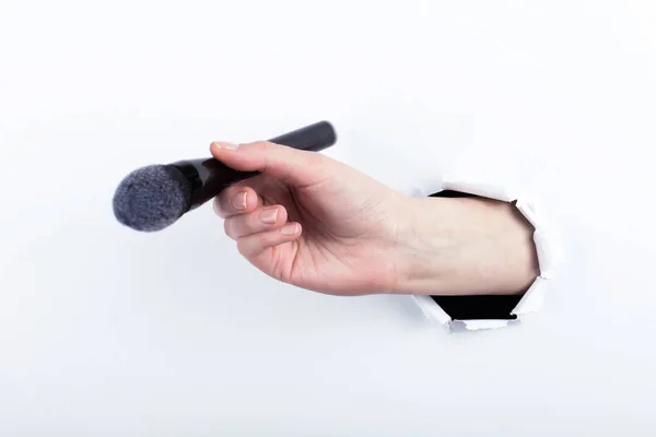 Female hand out of a hole in paper, holding a large powder brush. Isolate on white background.