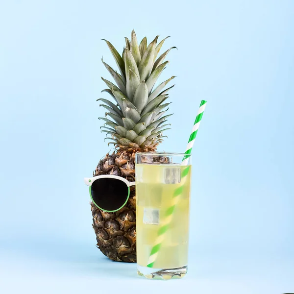 Ananas dans des lunettes de soleil et jus dans un verre, concept d'humeur estivale drôle, sur un fond bleu . — Photo
