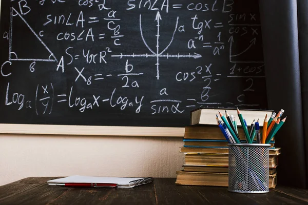 School desk in classroom, with books on background of chalk board with written formulas. Soncept Teacher's Day.