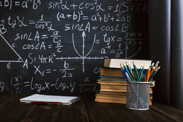 School desk in classroom, with books on background of chalk board with written formulas. Soncept Teacher's Day.