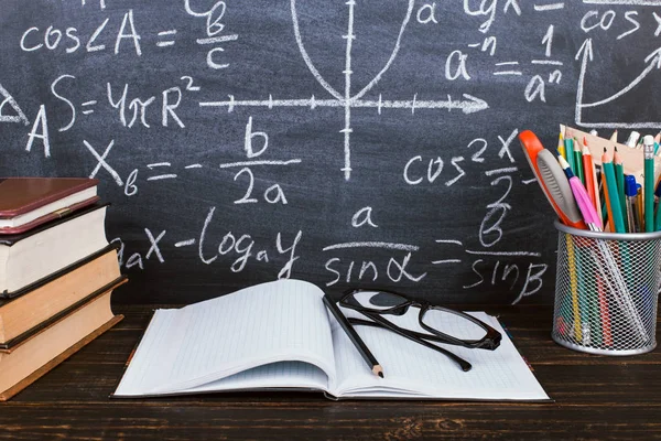 笔记本、玻璃杯和带铅笔的玻璃在黑板上，背景有公式。教师节概念与返校. — 图库照片