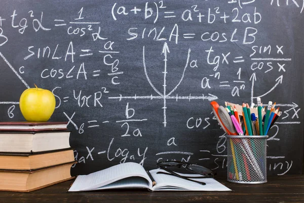 Notizbuch, Brille, Apfel und ein Glas mit Bleistiften auf Tafelhintergrund mit Formeln. Lehrertagskonzept und zurück zur Schule. — Stockfoto