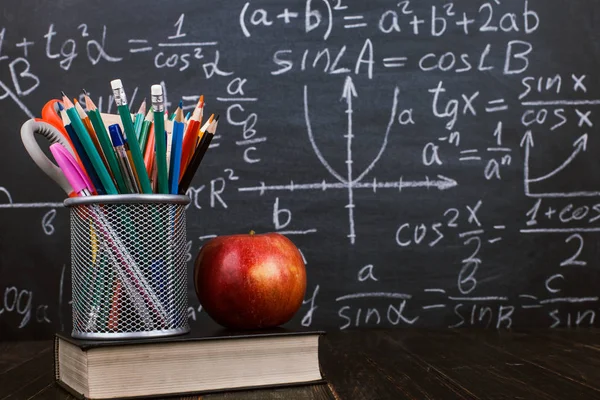 Libri, una mela, sta per penne su tavolo di legno, sullo sfondo di lavagna di gesso con formule. Concetto di giorno d'insegnante e ritorno a scuola . — Foto Stock