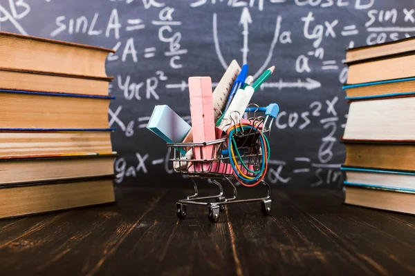 Shopping cart with school supplies, on the table with books against the background of a chalkboard. Concept back to school preparation.