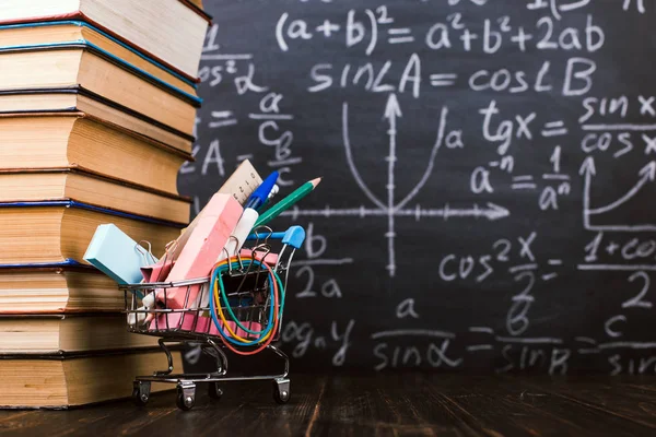 Okul malzemeleri ile alışveriş sepeti, bir tebeşir tahtası arka plan aleyhine kitaplar ile masada. Kavram geri okula hazırlık.