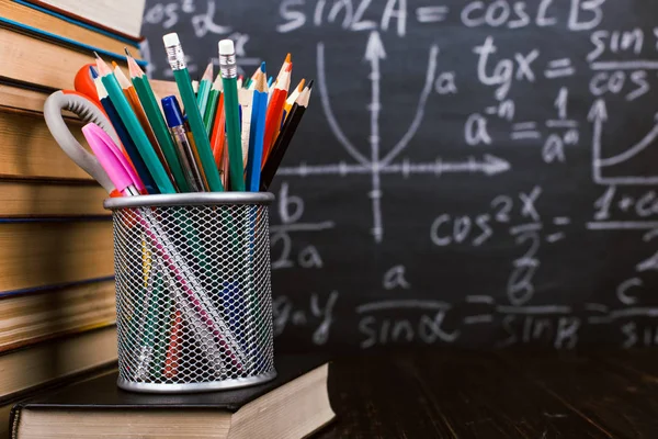 Böcker och stativ för pennor på ett träbord, mot bakgrund av en krita ombord med formler. Lärardagens koncept och tillbaka till skolan. — Stockfoto