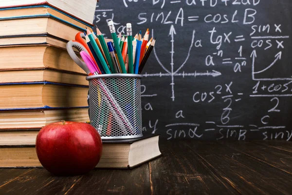 Los libros, una manzana, representan bolígrafos sobre una mesa de madera, sobre el fondo de una pizarra con fórmulas. Concepto del día del maestro y de vuelta a la escuela . — Foto de Stock