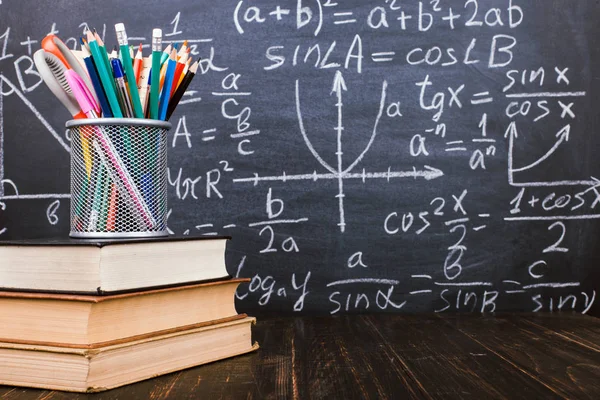 Boeken en staan voor pennen op een houten tafel, tegen de achtergrond van een krijtbord met formules. Teacher's Day concept en terug naar school. — Stockfoto