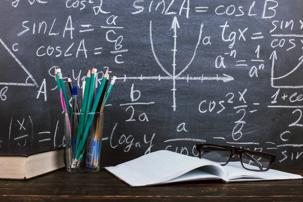 Caderno, óculos e um copo com lápis sobre fundo de quadro com fórmulas. Conceito do dia do professor e de volta à escola . — Fotografia de Stock