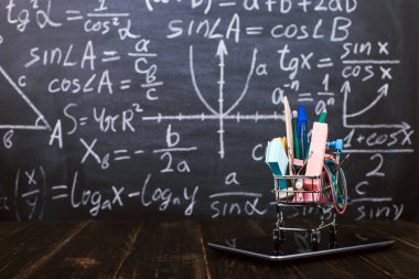 Arka plan tebeşir tahtasına karşı masada, tablette okul malzemeleri olan bir alışveriş arabası. Kavram geri okul hazırlık ve online alışveriş.