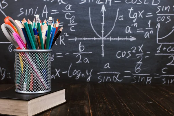 Libri e stand per penne su un tavolo di legno, sullo sfondo di una lavagna di gesso con formule. Concetto di giorno d'insegnante e ritorno a scuola . — Foto Stock