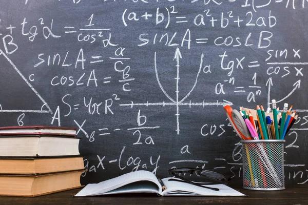 Bücher und Stifte auf einem Holztisch, vor dem Hintergrund einer Kreidetafel mit Formeln. Lehrertagskonzept und zurück zur Schule. — Stockfoto