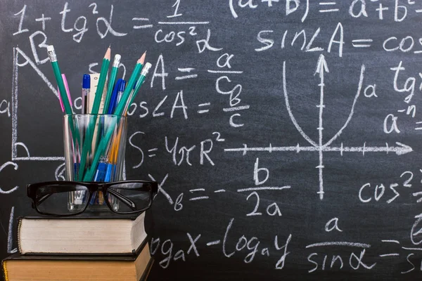 Libros y soportes para bolígrafos sobre una mesa de madera, sobre el fondo de una pizarra de tiza con fórmulas. Concepto del día del maestro y de vuelta a la escuela . — Foto de Stock