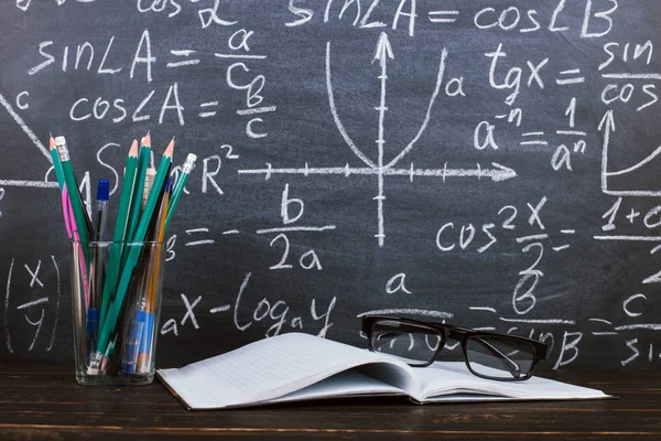 Poznámkový blok, brýle a sklo s tužkami v pozadí na tabuli se vzorci. Pojetí učitelova dne a zpět do školy. — Stock fotografie