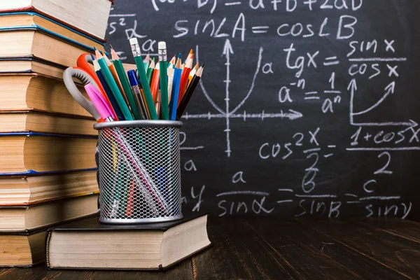 Libri e stand per penne su un tavolo di legno, sullo sfondo di una lavagna di gesso con formule. Concetto di giorno d'insegnante e ritorno a scuola . — Foto Stock