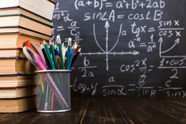 Böcker och stativ för pennor på ett träbord, mot bakgrund av en krita ombord med formler. Lärardagens koncept och tillbaka till skolan. — Stockfoto