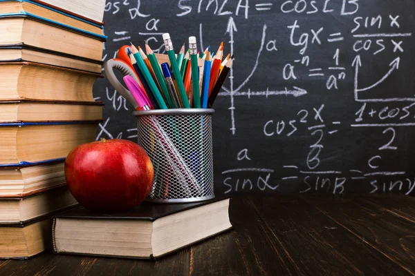 Livros, uma maçã, suporte para canetas em mesa de madeira, contra o fundo de tábua de giz com fórmulas. Conceito do dia do professor e de volta à escola . — Fotografia de Stock