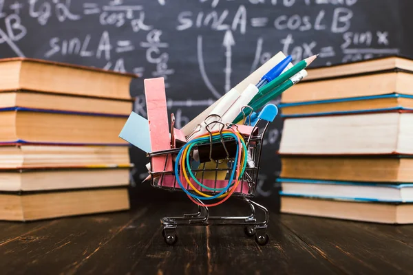 Shopping cart with school supplies, on the table with books against the background of a chalkboard. Concept back to school preparation.