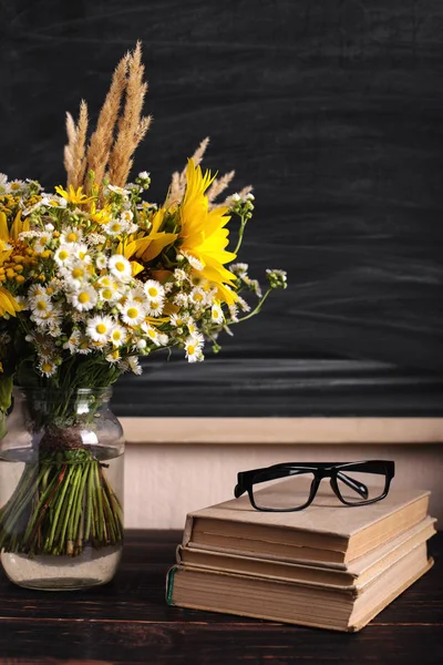Glasses teacher books and wildflowers bouquet on the table, on background blackboard with chalk. The concept of the teacher\'s day. Copy space.