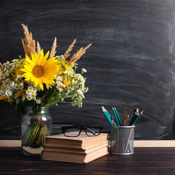 Glasses teacher books and wildflowers bouquet on the table, on background blackboard with chalk. The concept of the teacher\'s day. Copy space.