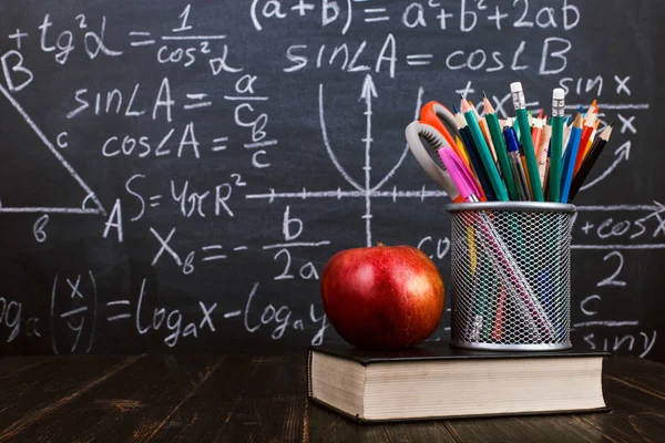 Books, an apple, stand for pens on wooden table, against the background of chalk board with formulas. Teacher's day concept and back to school.