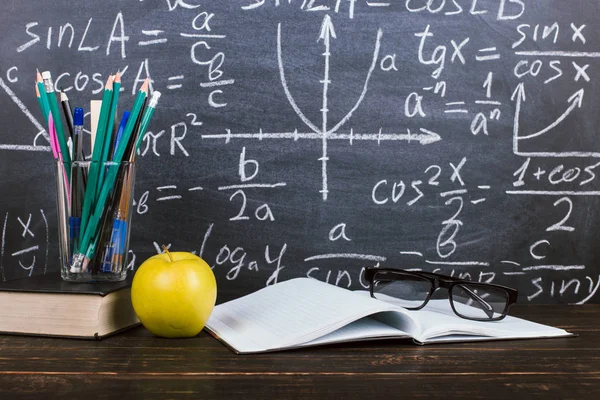 Notizbuch, Brille, Apfel und ein Glas mit Bleistiften auf Tafelhintergrund mit Formeln. Lehrertagskonzept und zurück zur Schule. — Stockfoto