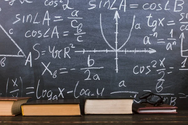 Boeken op een houten tafel, tegen de achtergrond van een krijtbord met formules. Teacher's Day concept en terug naar school. — Stockfoto