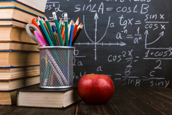 Libri, una mela, sta per penne su tavolo di legno, sullo sfondo di lavagna di gesso con formule. Concetto di giorno d'insegnante e ritorno a scuola . — Foto Stock