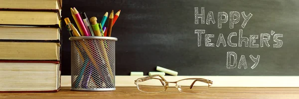 Glasses teacher books and a stand with pencils on the table, on the background of a blackboard with chalk. The concept of the teacher\'s day. Copy space.