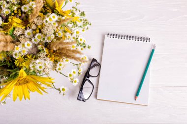 Beyaz bir masaüzerinde çiçek, defter ve kalem buketi. Kopyalama alanı.