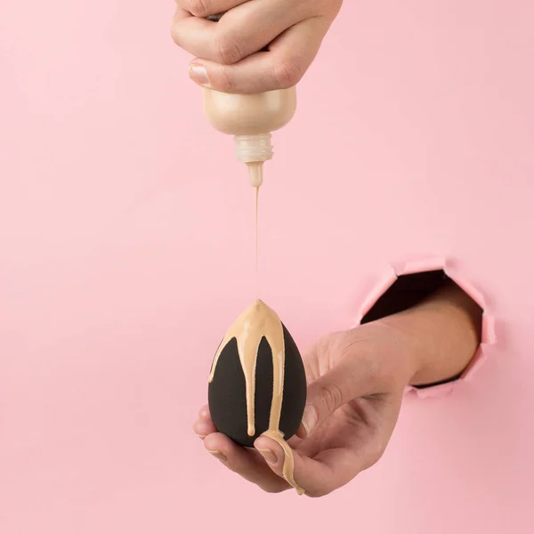 Girl hand holds a beauty blender in foundation, concealer from a hole in a pink background. Makeup artist concept, copy space. — Stock Photo, Image