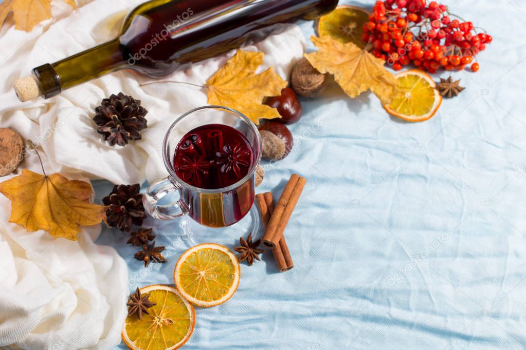 A cup of mulled wine with spices, bottle, dry leaves and oranges on table. Autumn mood, method to keep warm in the cold, copy space, morning light.
