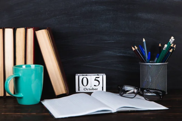 Kitaplar, takvim, defterler, kalemler, bardaklar ve bir fincan kahve, bir karatahtanın arka planında. Öğretmenler Günü için konsept. Boşluğu kopyala.