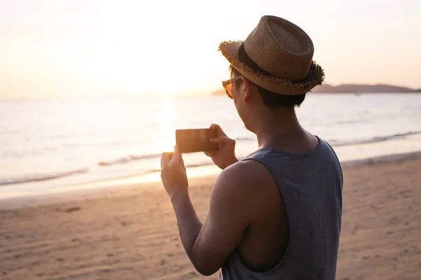 Silhueta de volta do jovem mochileiro de viagem masculino com chapéu tirando fotos da paisagem da praia de verão contra o pôr do sol - imagem de viagem momento capturado conceito . — Fotografia de Stock