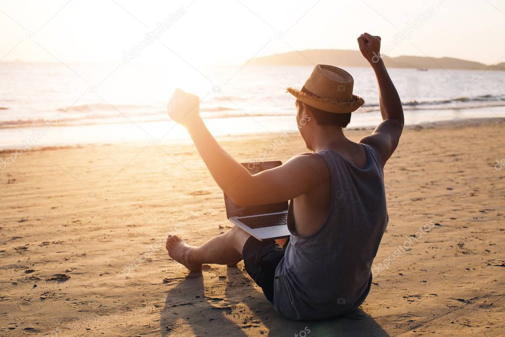 Young male businessman putting hands up for success on tropical summer beach - it indicates freedom such as financial freedom or remote work concept.