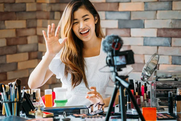 Sonriente mujer asiática agitando la mano en la cámara en la mesa con cosméticos — Foto de Stock