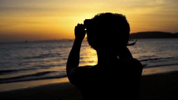 Silhouette posteriore di giovane viaggiatore maschile zaino in spalla con cappello scattare foto di scenario spiaggia estiva contro il tramonto - foto di viaggio momento catturato concetto — Video Stock