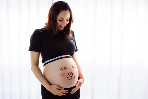 Mladá žena s nakreslenými obličeji úsměv na těhotné bříško v ložnici — Stock fotografie