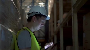 Güvenlik kasketi olan genç bir depo çalışanı sipariş detaylarını envanter odasında dijital tabletle kontrol ediyor.