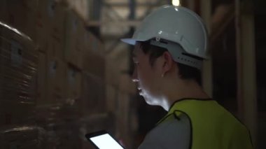 Güvenlik kasketi olan genç bir depo çalışanı elinde dijital tabletle envanter odasında yürüyor.