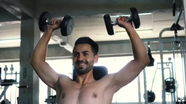 Мускулистый сильный молодой человек, поднимающий плечи с гантелями в тренажерном зале — стоковое видео