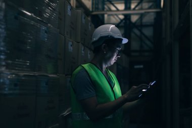 Güvenlik kasketi olan genç bir depo çalışanı sipariş detaylarını envanter odasında dijital tabletle kontrol ediyor.