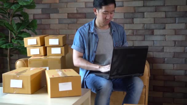 Азиатский предприниматель-мужчина, использующий ноутбук с пакетами коробок дома — стоковое видео