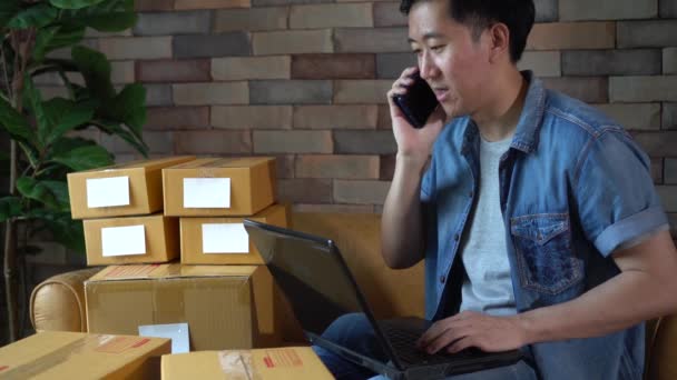 Asian male business entrepreneur using laptop and phone with packs of boxes at home — Stock Video