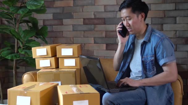 Азиатский предприниматель-мужчина, использующий ноутбук и телефон с пакетами коробок дома — стоковое видео