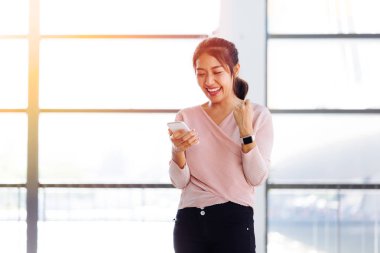 Young Asian girl looking at her mobile phone and get excited inside the building clipart