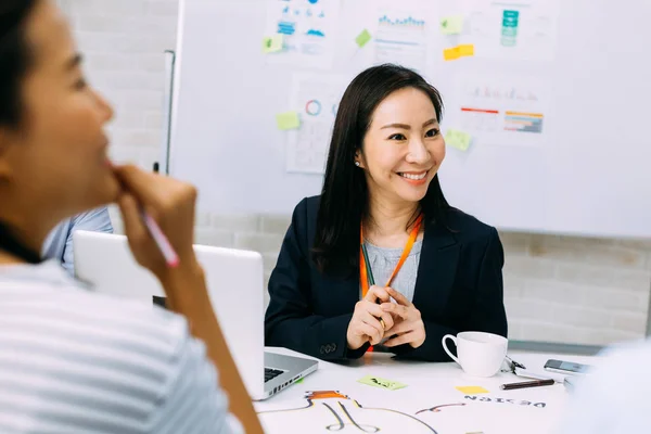 Ασιάτισσα ώριμη επιχειρηματίας χαμογελώντας και κοιτάζοντας άλλους επιχειρηματίες, ενώ κάθεται στη συνάντηση — Φωτογραφία Αρχείου