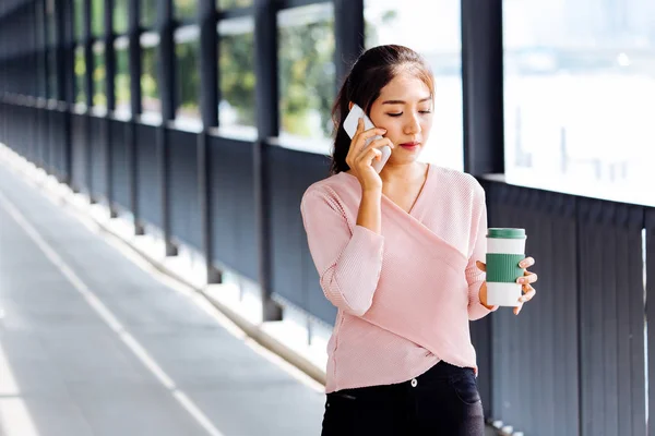Asian woman speaking on smartphone and walking with coffee in hand