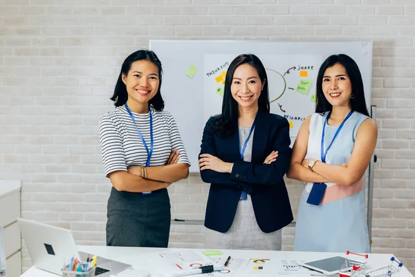 Sonrientes mujeres de negocios jóvenes asiáticas en ropa casual de pie en línea con los brazos doblados en la sala de reuniones — Foto de Stock